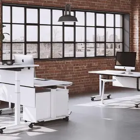 Modernes Büro mit höhenverstellbaren Schreibtischen, großen Fenstern und industriellem Loft-Design.