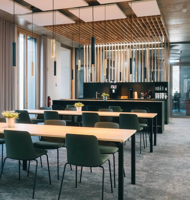 Moderner Pausenraum mit Holztischen, grünen Stühlen und offener Küche im Bürogebäude.