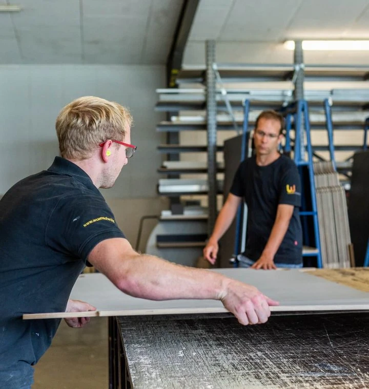 Zwei Handwerker schneiden in einer modernen Werkstatt gemeinsam eine große Holzplatte zurecht.